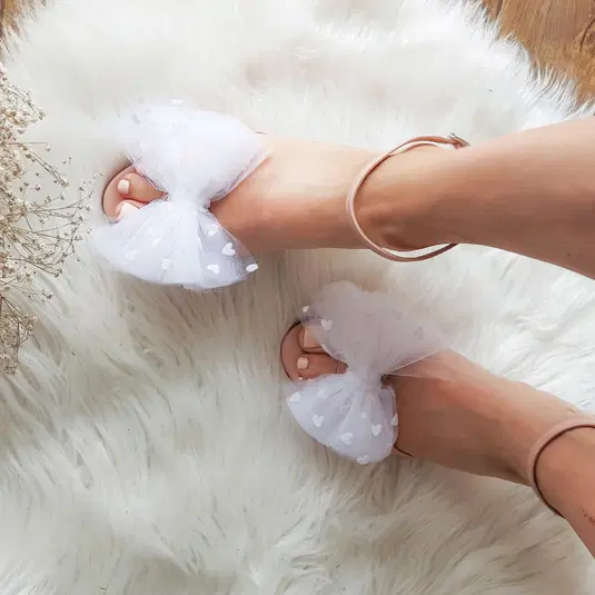  Białe sandały z delikatną koronkową kokardą na beżowym dywanie, moda ślubna, eleganckie obuwie damskie. 