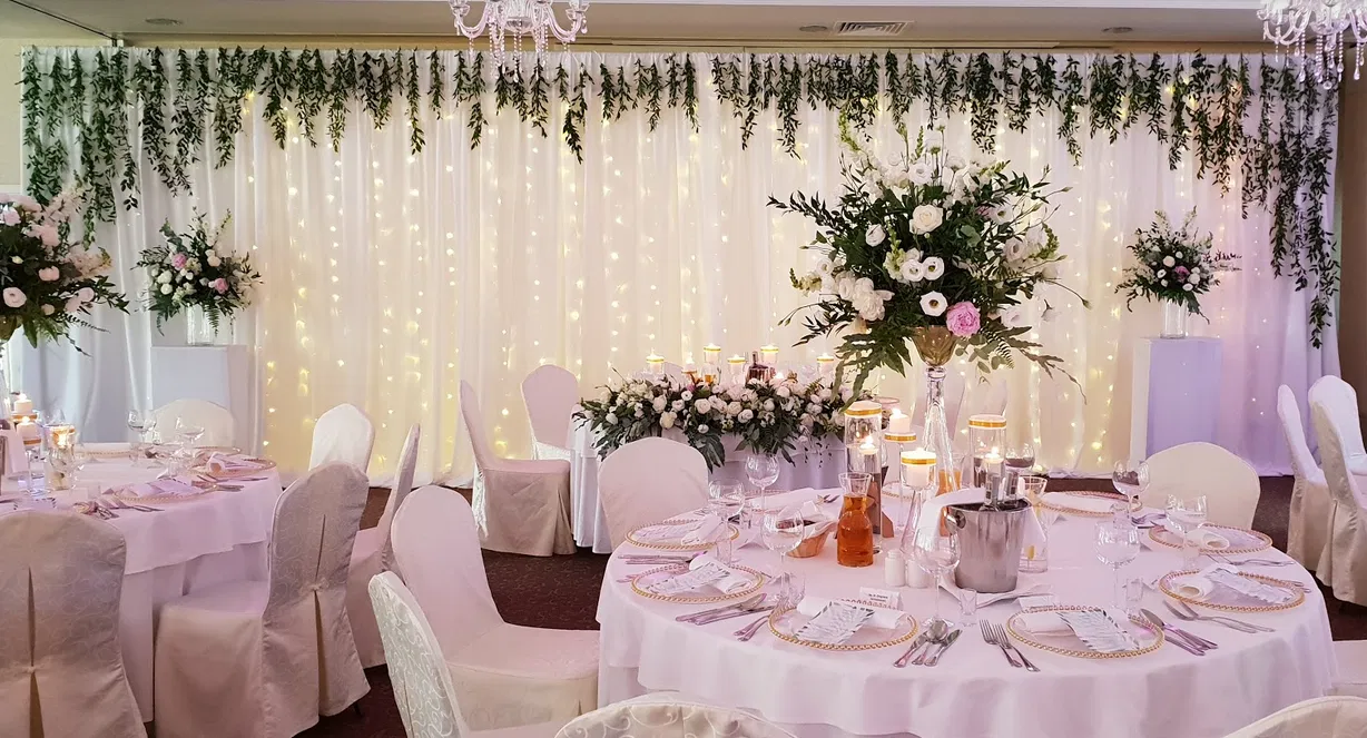 Elegancka dekoracja sali weselnej z białymi obrusami, kwiatami, świecami i świetlną kurtyną.