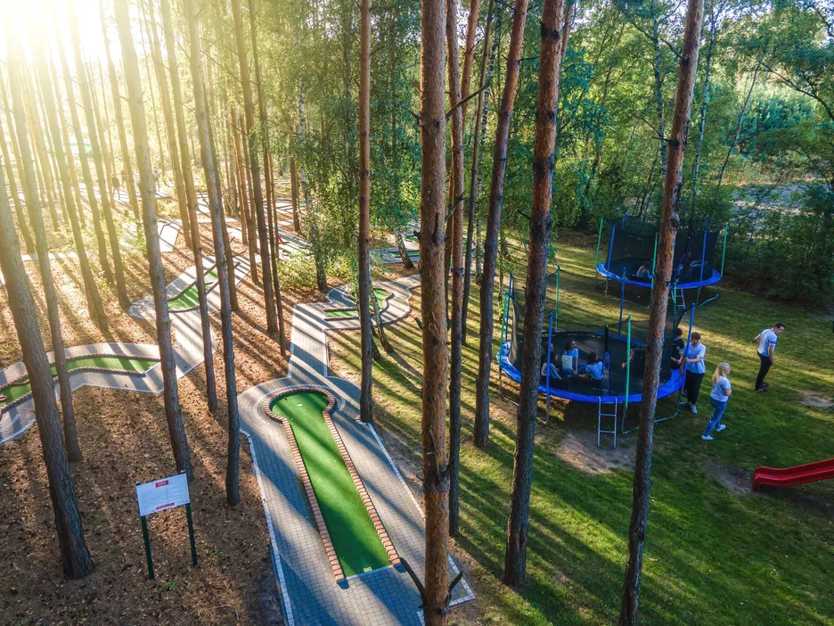 Leśny park rozrywki z mini-golfem i trampolinami otoczony drzewami w promieniach słońca.
