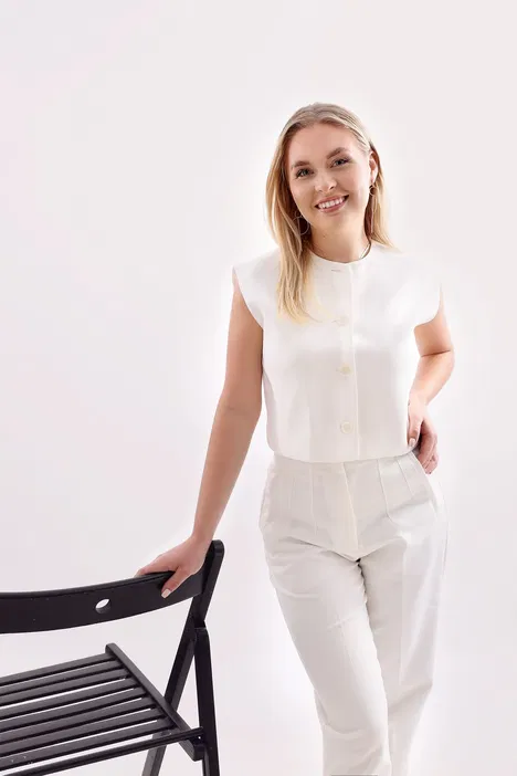 Blondynka w białym eleganckim stroju na tle jasnego wnętrza, trzymająca czarne krzesło.