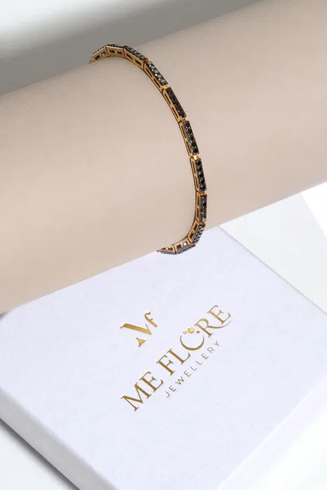 Złota bransoletka z czarnymi kamieniami na eleganckim beżowym tle, obok opakowania z logo Me Flore Jewellery.