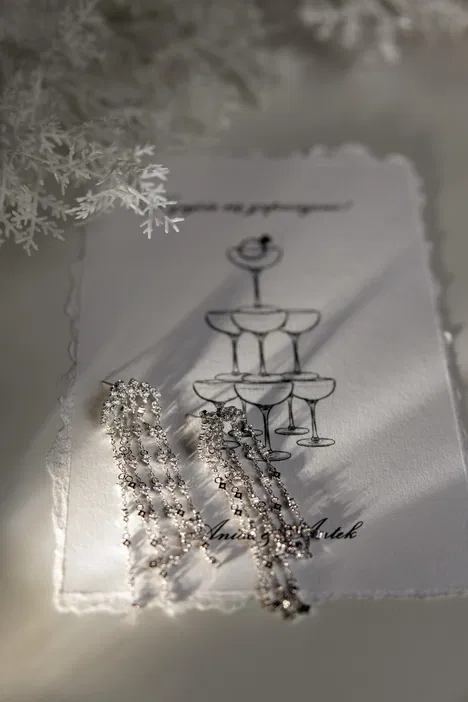Wiszące kolczyki na tle eleganckiego zaproszenia z rysunkiem kieliszków do szampana, dekorowane białymi gałązkami, nawiązujące do uroczystego wydarzenia.