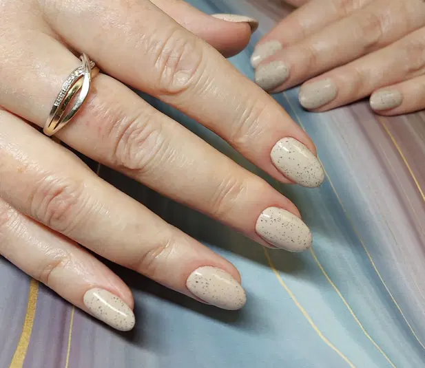  Dłonie z eleganckim, beżowym manicure z brokatowym wzorem na pastelowym tle. Obrączka na jednym z palców. Idealny przykład stylowego, subtelnego zdobienia paznokci. 
