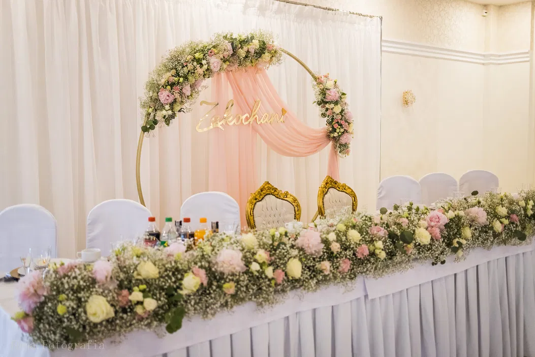 Elegancka dekoracja stołu weselnego z kwiatowym łukiem i złotymi krzesłami, różowe i białe kwiaty, napis Zakochani, sala weselna.