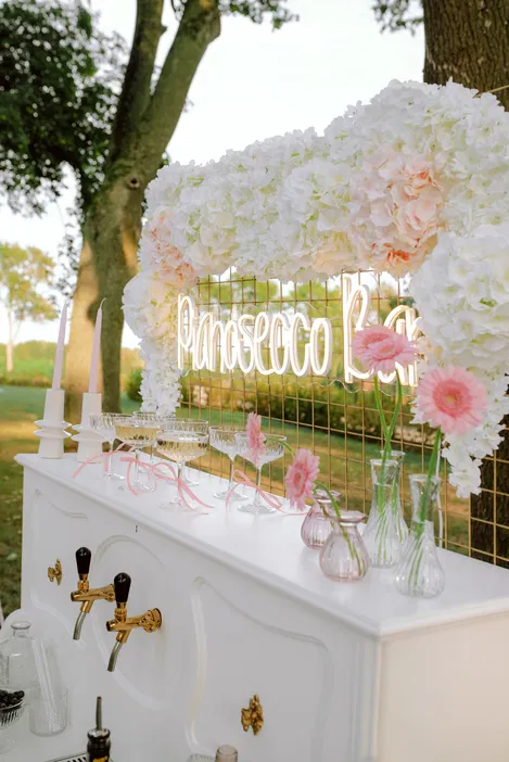 Kwiatowy neonowy prosecco bar na świeżym powietrzu z dekoracjami z białych i różowych kwiatów, eleganckie kieliszki do szampana, idealne na letnie przyjęcia.