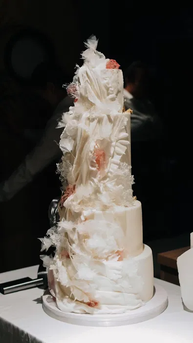 Tort weselny o eleganckim i artystycznym designie w jasnych kolorach, ozdobiony delikatnymi dekoracjami z cukru, na ciemnym tle.