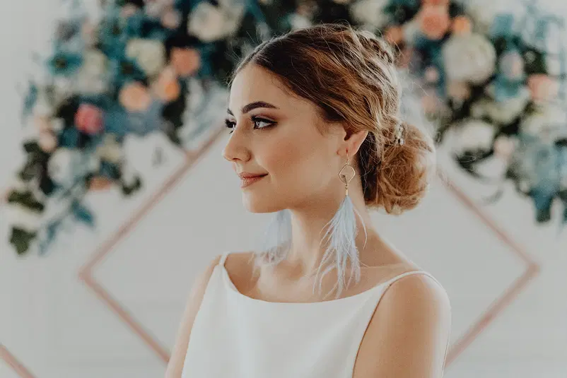  Brunetka w białej sukni z upiętymi włosami i niebieskimi piórkami w uszach, na tle kolorowych kwiatów. 