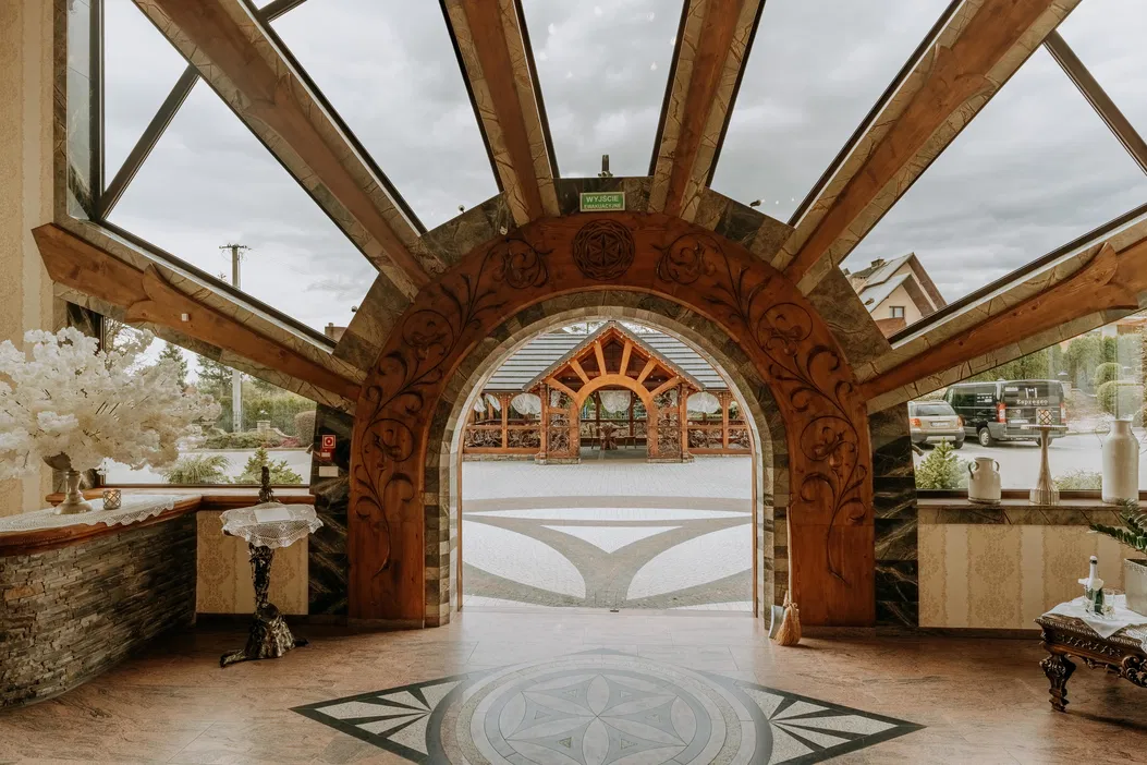 Wnętrze eleganckiego budynku z ozdobnym drewnianym łukiem, widok na zewnątrz z zadaszonym patio, dekoracyjne elementy i nowoczesna architektura.
