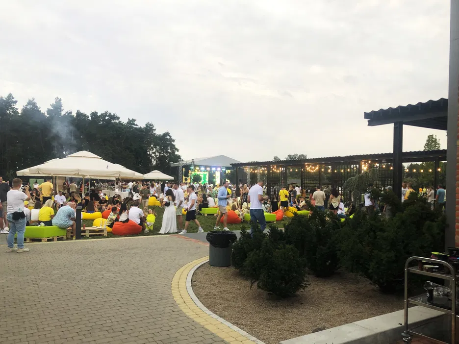 Outdoor event with large crowd, colorful seating, and string lights; people gathered for a festival or celebration near a wooded area.