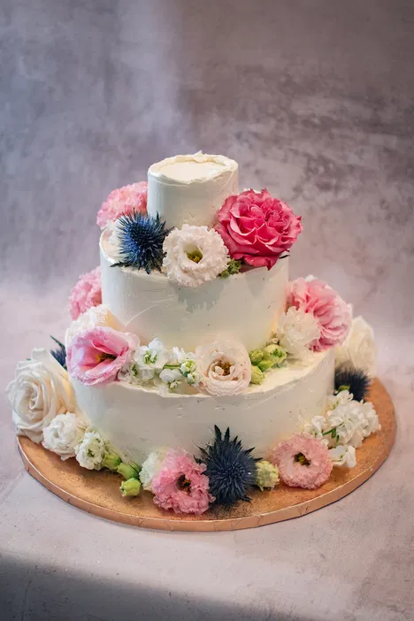 Tort weselny z trzema poziomami ozdobiony świeżymi kwiatami w pastelowych kolorach na ciepłym tle.