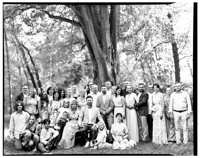  Grupa ludzi na zewnątrz w parku, zdjęcie czarno-białe, uroczystość rodzinna, eleganckie stroje, duże drzewo w tle, fotografia grupowa w plenerze. 