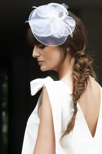 Kobieta w eleganckiej, białej sukience z warkoczem i fascynatorem na głowie, profil zbliżony, ciemne tło. 