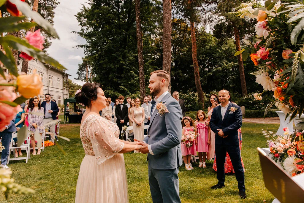 Para młoda podczas ceremonii ślubnej na świeżym powietrzu w ogrodzie, otoczona gośćmi i kolorowymi kwiatami.