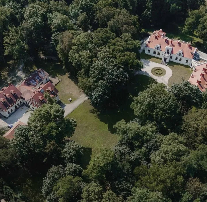 Widok z lotu ptaka na zabytkowy pałac otoczony zielenią, z czerwonymi dachami i starannie utrzymanym parkiem na tle gęstych drzew.