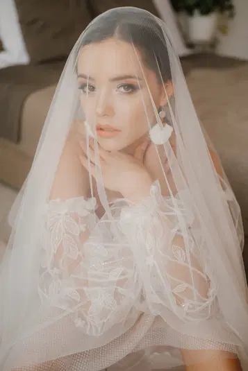  Brunetka w koronkowej sukni ślubnej i welonie, z długimi kolczykami, siedząca na kanapie w naturalnym oświetleniu. 
