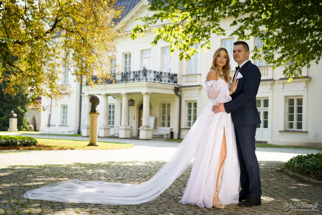 Para młoda w eleganckich strojach ślubnych stoi przed stylowym, białym pałacem otoczonym zielenią, idealne miejsce na romantyczną sesję fotograficzną na świeżym powietrzu.