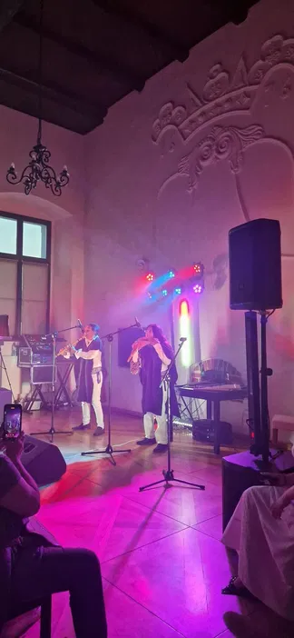 Muzycy grający na tradycyjnych instrumentach w oświetlonej sali koncertowej, z widocznymi mikrofonami i kolorowymi światłami na scenie.