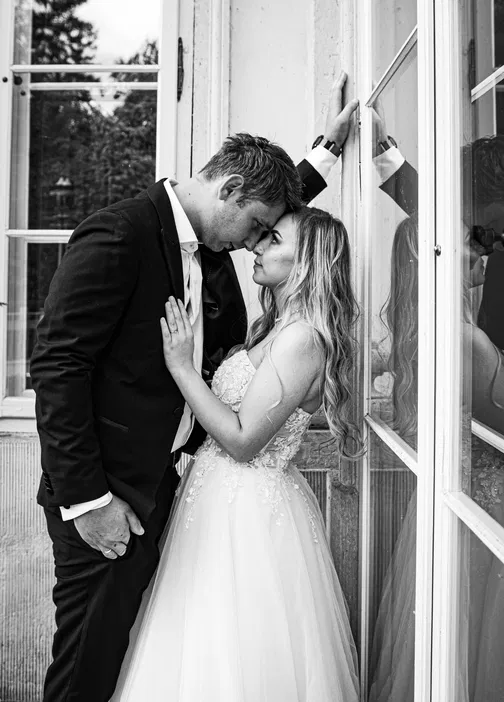 Romantyczna czarno-biała fotografia ślubna - para młoda przy oknie w czułym uścisku, elegancki garnitur i koronkowa suknia, intymny moment fotografowany w stylu vintage.