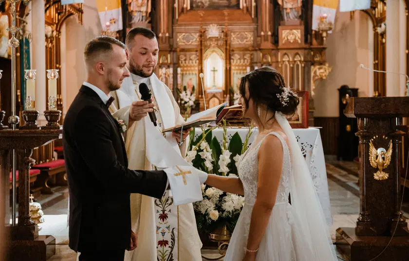  Ceremonia ślubna w kościele, para młoda wymienia przysięgi ślubne, ksiądz trzyma mikrofon i książkę liturgiczną, dekoracje z kwiatów i krzyż w tle. 