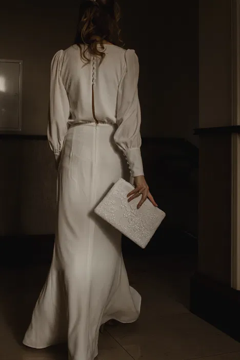 Kobieta w eleganckiej, długiej białej sukni z bufkami, trzymająca wyszywaną kopertówkę, widziana od tyłu.