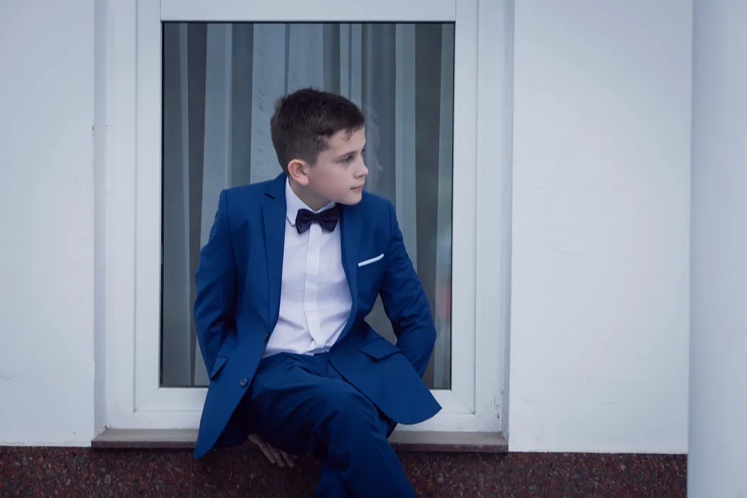 Chłopiec w eleganckim niebieskim garniturze siedzący na parapecie okna.
