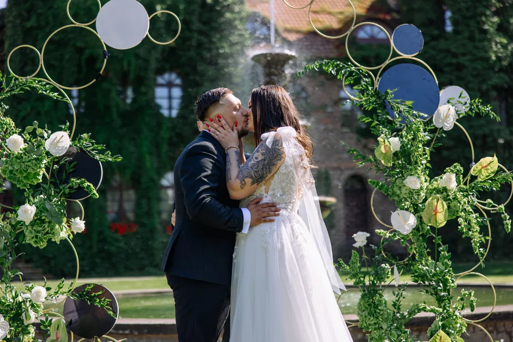 Para młoda całuje się na tle zielonego ogrodu, dekoracje weselne z kwiatami i okręgami.