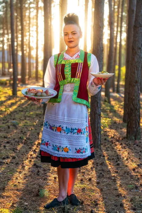 Kobieta w tradycyjnym stroju ludowym podająca potrawy na talerzach w lesie o zachodzie słońca.