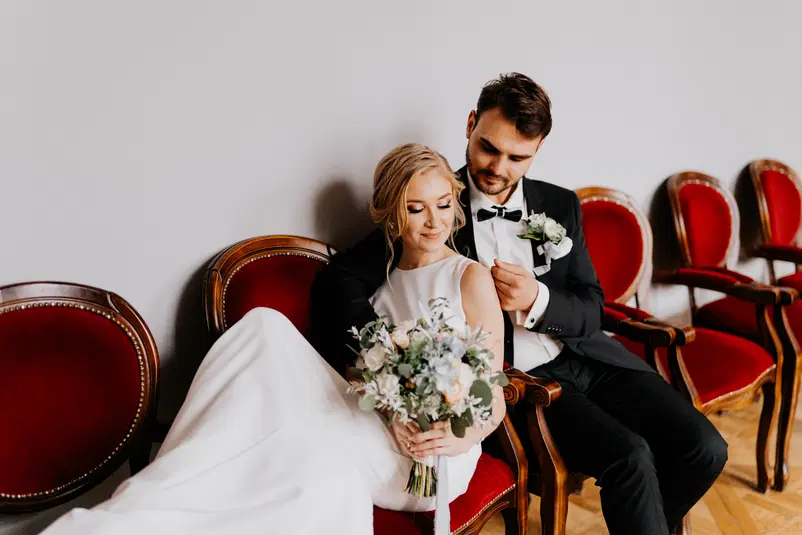 Para młoda siedząca na czerwonych krzesłach, trzymająca bukiet kwiatów, elegancko ubrana na ślubie. 