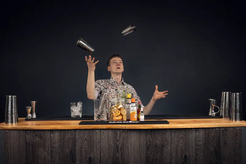  Barman żongluje shakerami w eleganckim barze, otoczony akcesoriami barmańskimi i składnikami do drinków. 