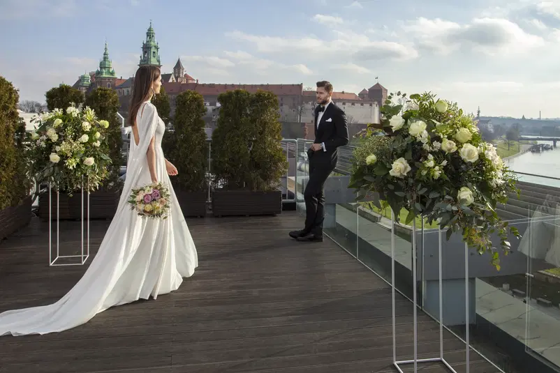 Para ślubna na tarasie z widokiem na Wawel, elegancka panna młoda w białej sukni z bukietem, stylowy pan młody w garniturze, romantyczna sceneria z kwiatowymi dekoracjami. 