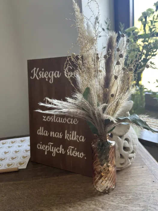 Dekoracja stołu z suszonymi kwiatami w brązowym wazonie i drewnianą tablicą z napisem Księga Gości, zachęcająca do zostawienia ciepłych słów.