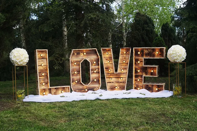  Duże drewniane litery z oświetleniem, tworzące słowo LOVE, stojące na tle zielonych drzew, ozdobione kwiatowymi dekoracjami z białych róż. 