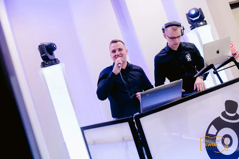  DJ na imprezie, prowadzenie muzyki, stanowisko DJ-skie, sprzęt audio, atmosfera wydarzenia, oświetlenie sceniczne. 