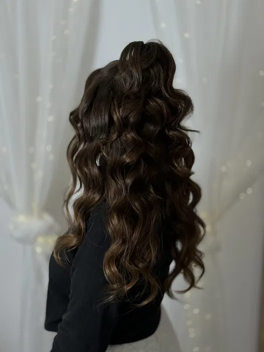 Kobieta z długimi, ciemnobrązowymi, falującymi włosami upiętymi w elegancki kok, stojąca przed białą zasłoną.
