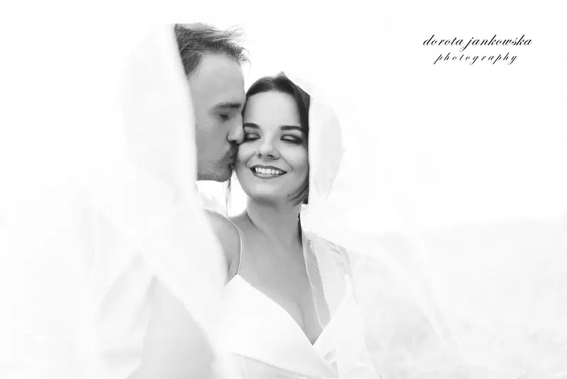  Czarno-białe zdjęcie romantyczne pary, mężczyzna całuje kobietę w policzek, oboje uśmiechnięci, osłonięci welonem, fotografia ślubna. 