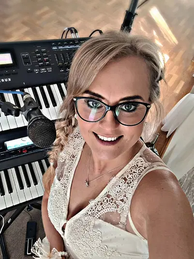  Osoba w okularach przed mikrofonem i klawiaturą elektroniczną, uśmiechnięta i ubrana w elegancką białą sukienkę z koronkowymi zdobieniami. 