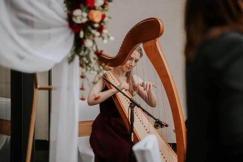  Harfistka grająca na harfie podczas ceremonii, dekoracje kwiatowe w tle, elegancka atmosfera. 