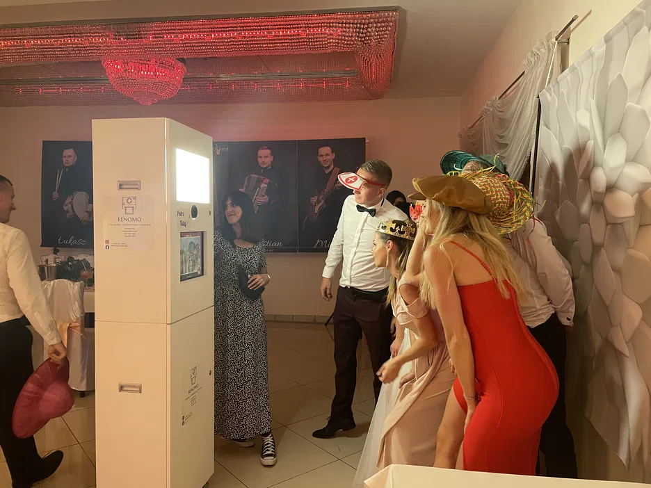 Ludzie pozujący przy fotobudce na imprezie, w kolorowych przebraniach, czapki i rekwizyty na głowach, w eleganckiej sali z czerwonym oświetleniem i dekoracjami.