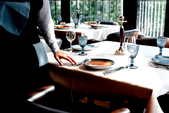  Elegancko nakryty stół w restauracji z zastawą, kieliszkami i sztućcami, obsługiwany przez kelnera. 