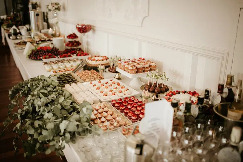  Stół z deserami na weselu, pełen ciast, babeczek, owoców i dekoracji z eukaliptusa, elegancki wystrój wnętrza, stoły weselne, słodki bufet. 