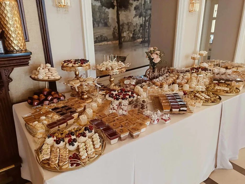 Stół deserowy na przyjęciu z różnorodnymi ciastami, tortami i słodkościami, ozdobiony eleganckimi paterami i dekoracjami, na tle eleganckiego pomieszczenia.