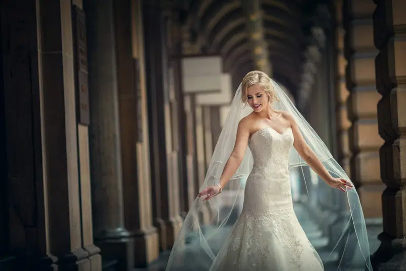  Piękna panna młoda w białej sukni ślubnej z welonem spaceruje po eleganckim arkadach. 