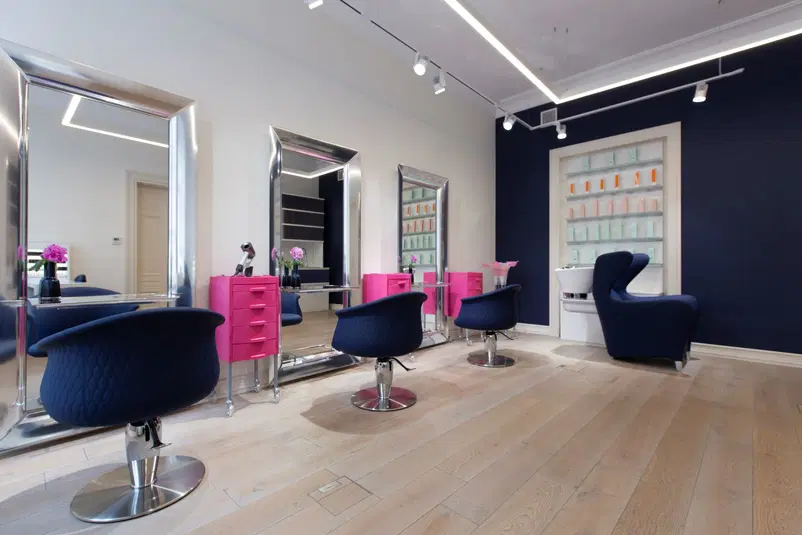  Wnętrze nowoczesnego salonu fryzjerskiego z eleganckimi krzesłami i różowymi szafkami, duże lustra, drewniana podłoga, ciemne ściany i półki z kosmetykami do pielęgnacji włosów. 