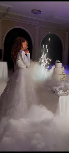  Panna młoda w białej sukni ślubnej stoi obok tortu weselnego z zimnymi ogniami, w otoczeniu dymu na eleganckiej sali. 