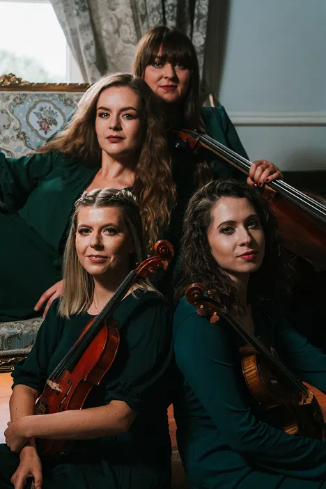 Cztery kobiety grające na instrumentach muzycznych, w eleganckich zielonych strojach, siedzące w klasycznym wnętrzu, artystyczna sesja fotograficzna kwartetu smyczkowego.