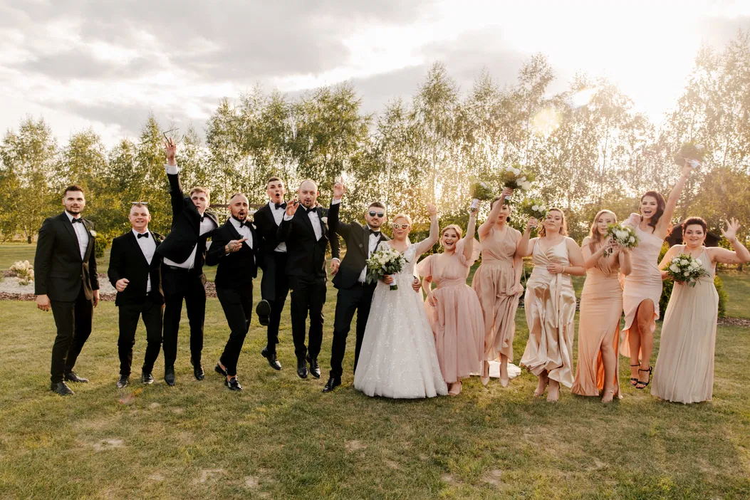 Grupa weselna pozująca w parku, uśmiechnięci goście w eleganckich strojach, panna młoda w białej sukni trzyma bukiet, naturalne oświetlenie letniego zachodu słońca.