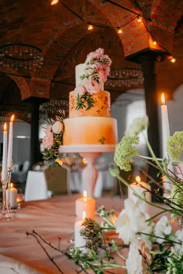  Trzypiętrowy tort weselny ozdobiony kwiatami i świecami w eleganckiej scenerii salonu z ceglanym sklepieniem. 