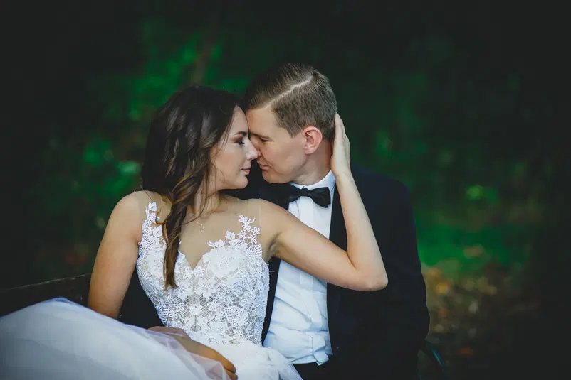  Para młoda siedząca na ławce w parku, ubrana w eleganckie stroje ślubne, patrząca sobie w oczy, romantyczna atmosfera. 