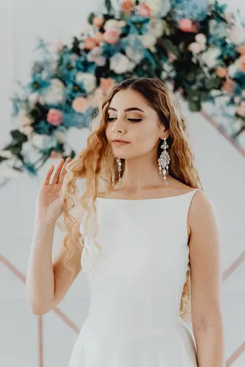  Młoda kobieta w białej sukience z długimi, falowanymi włosami i ozdobnymi kolczykami, stojąca przed kolorową, kwiatową aranżacją. 