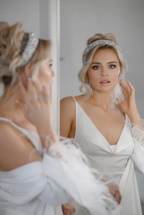 Blondynka w eleganckiej białej sukni ślubnej, patrząca w lustro, z delikatnym makijażem i ozdobną opaską na włosach.